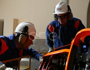 Due Operai, con casco, occhiali protettivi e cuffie antirumore, in sala macchine controllano il funzionamento dell'eccitatrice di una turbina